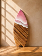 Wooden surfboard-shaped resin cheeseboard with a pink and white resin waves  on a beige wall.