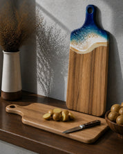 Camphor laurel cutting board with potatoes and a knife on a wooden surface, next to the same shape cutting board with a decorative resin handle in the beach colourway