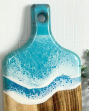 Wooden cutting board with a light blue and white resin wave design on a tiled wall background