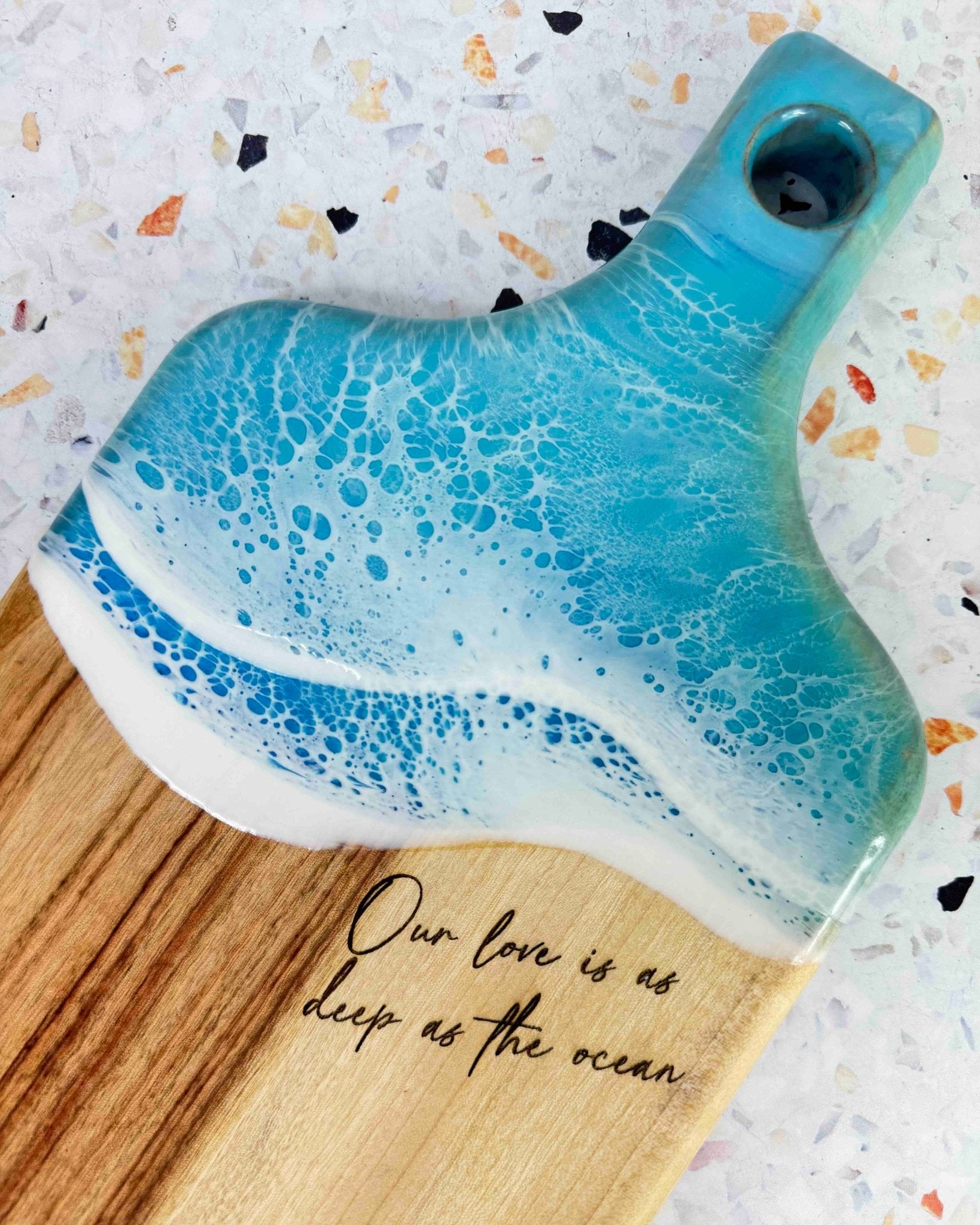 Decorative resin cheese board with light blue and white wave design and engraved text on a speckled surface. The engraving reads "our love is as deep as the ocean"