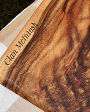 A close-up of a personalized burl wood plaque or board with the engraved name 'Clan McIntosh' on a beige and white checkered fabric surface.
