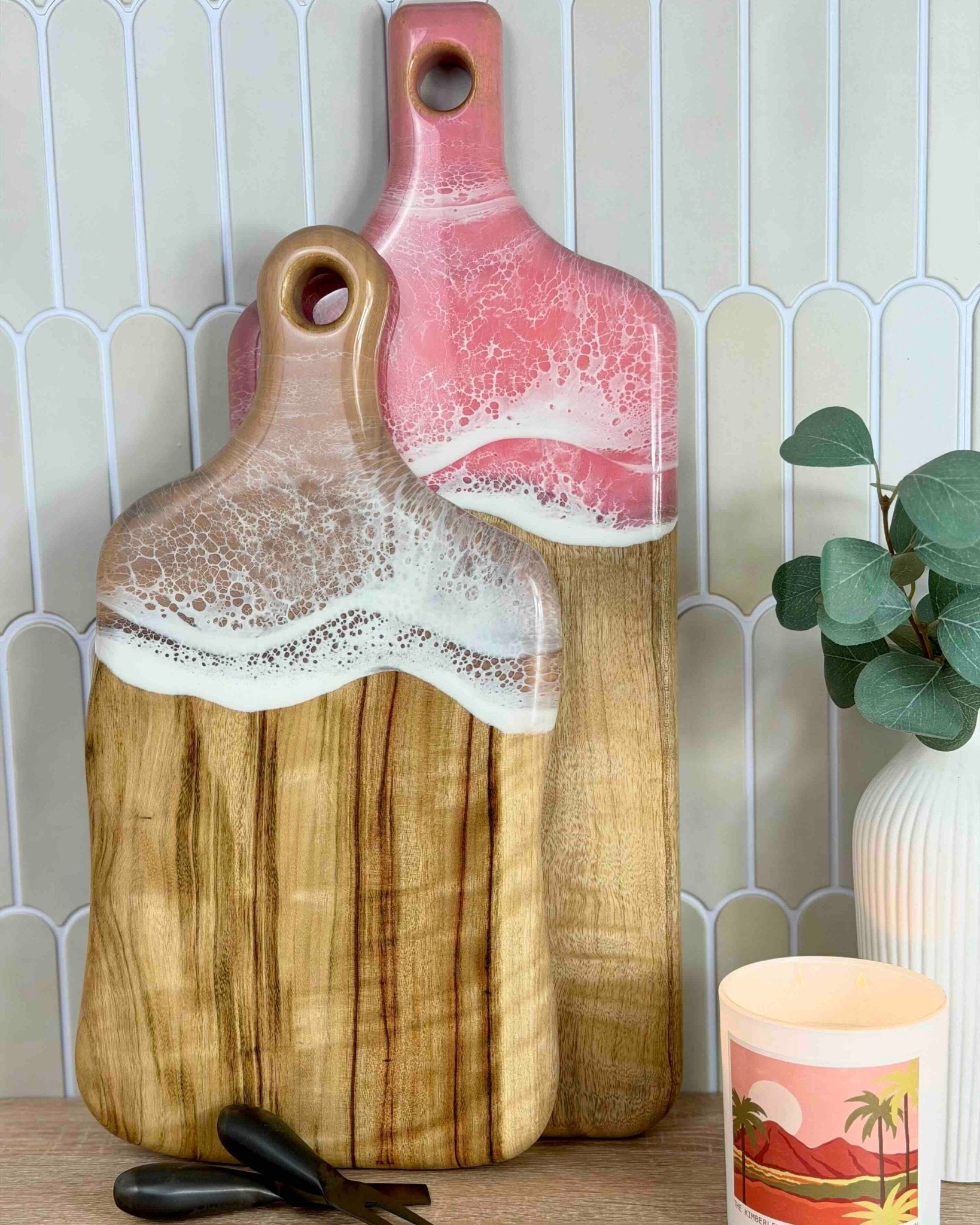 Two wooden cutting boards, one rectangle paddle, one medium paddle, with pink and brown handles on a kitchen counter.