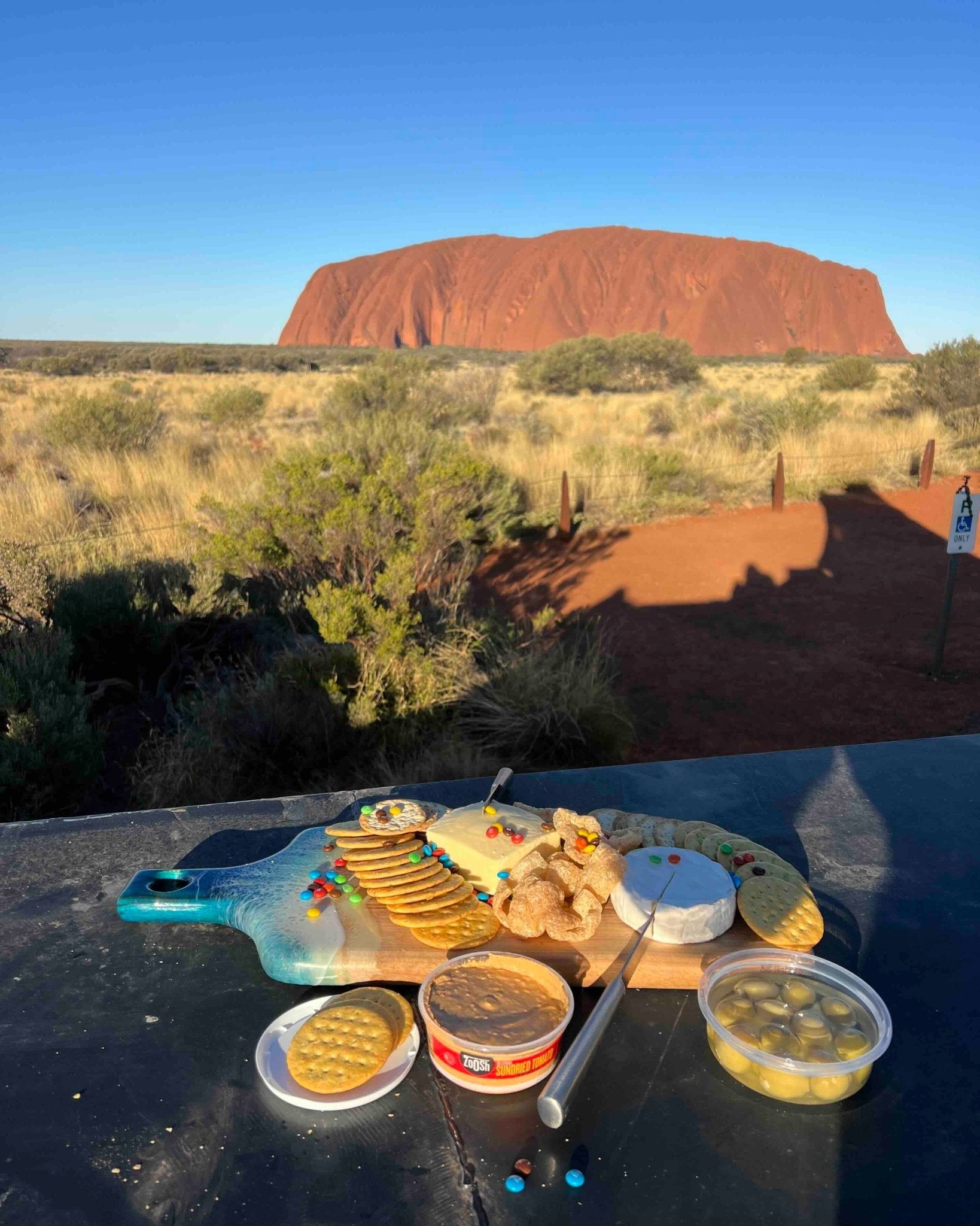 A picnic-style food spread arranged on a car canopy roof in a front of uluru, australia,  under a clear blue sky. The food includes crackers, cheese, bread, and small containers of snacks.