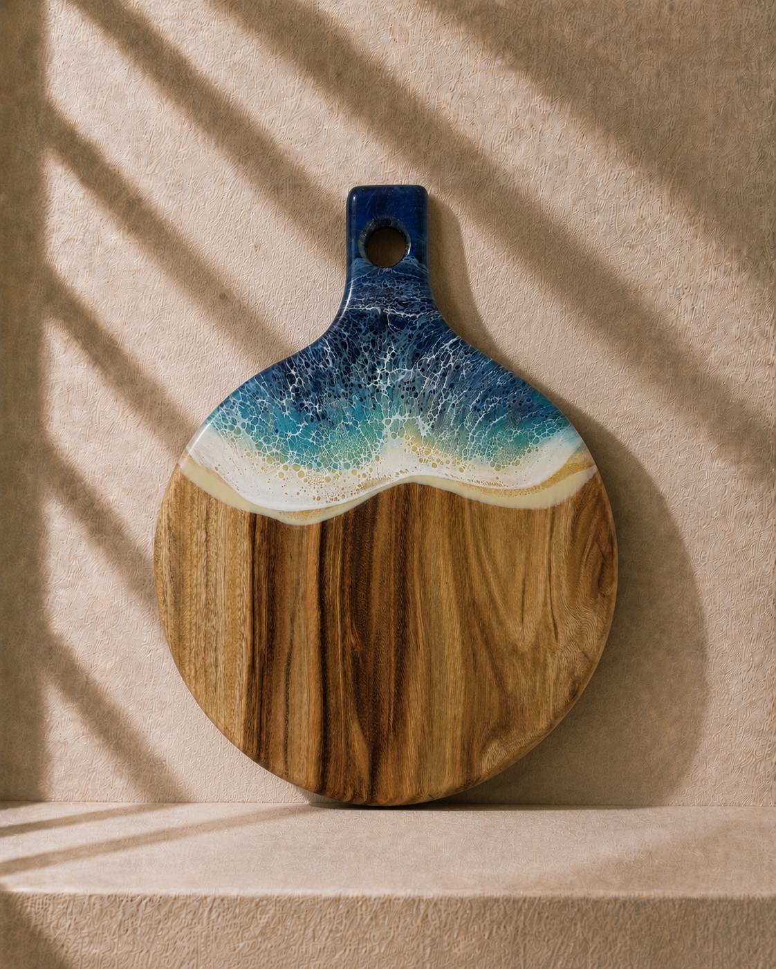 A round wooden serving board with a handle, featuring a wave design in blue, teal, and white above a natural wood grain base, displayed against a textured beige wall with soft shadow overlays.