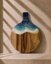 A round wooden serving board with a handle, featuring a wave design in blue, teal, and white above a natural wood grain base, displayed against a textured beige wall with soft shadow overlays.