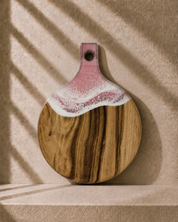 Round wooden serving board with a pink and white resin covered handle, displayed on a textured beige surface with soft window shadow overlays.