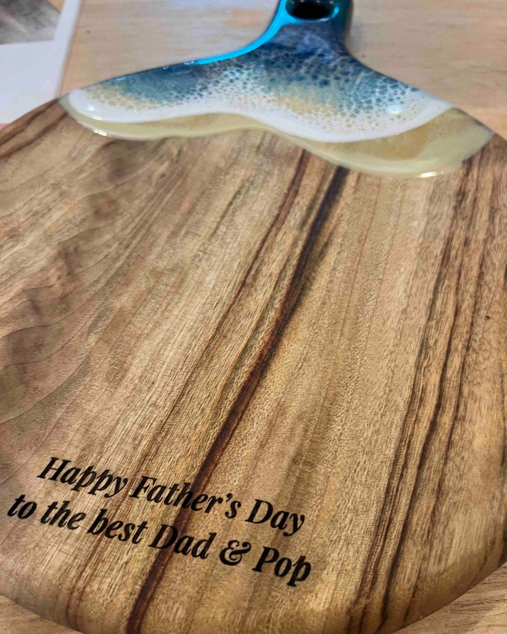 A round paddle board in the beach coloured resin showing the free engraving option saying "happy 70th birthday"