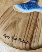 A round paddle board in the beach coloured resin showing the free engraving option saying "happy 70th birthday"