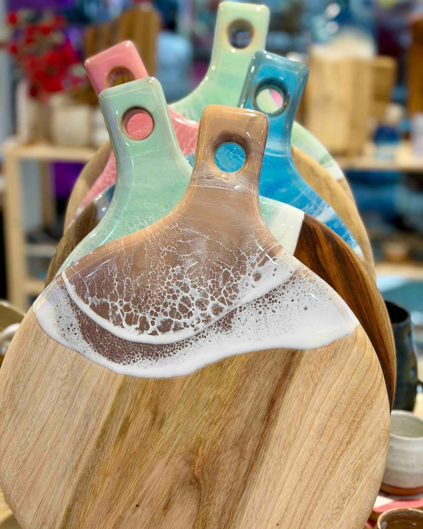 A close-up of a wooden serving board with colourful resin paddle handles in mint green, blue, and pink, set against a softly blurred background.