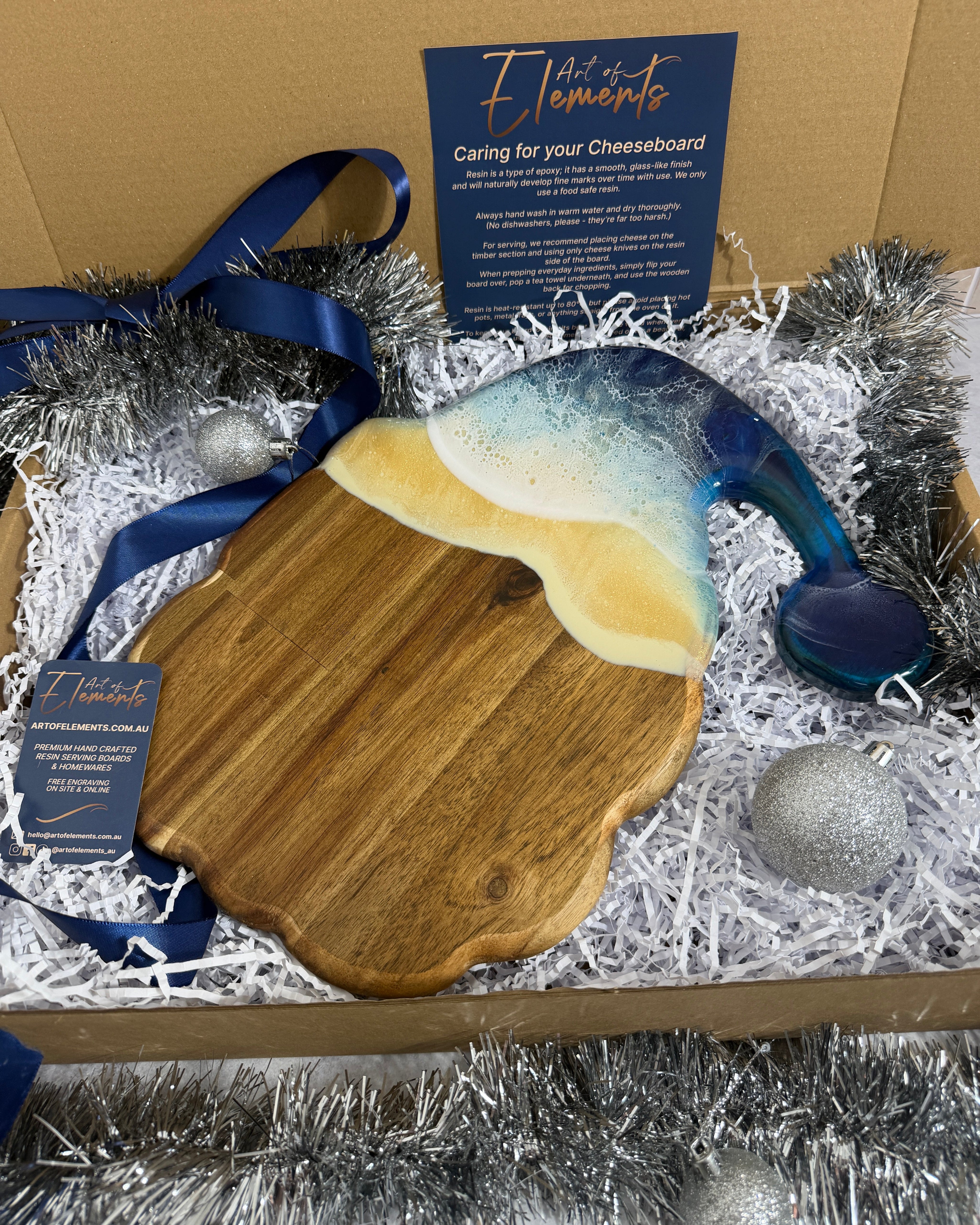 A wooden cheeseboard shaped as a santa head, with a glossy resin in beach colouring presented inside a gift box with shredded paper, silver tinsel, ornaments, and blue ribbons. Two blue brand cards are visible.