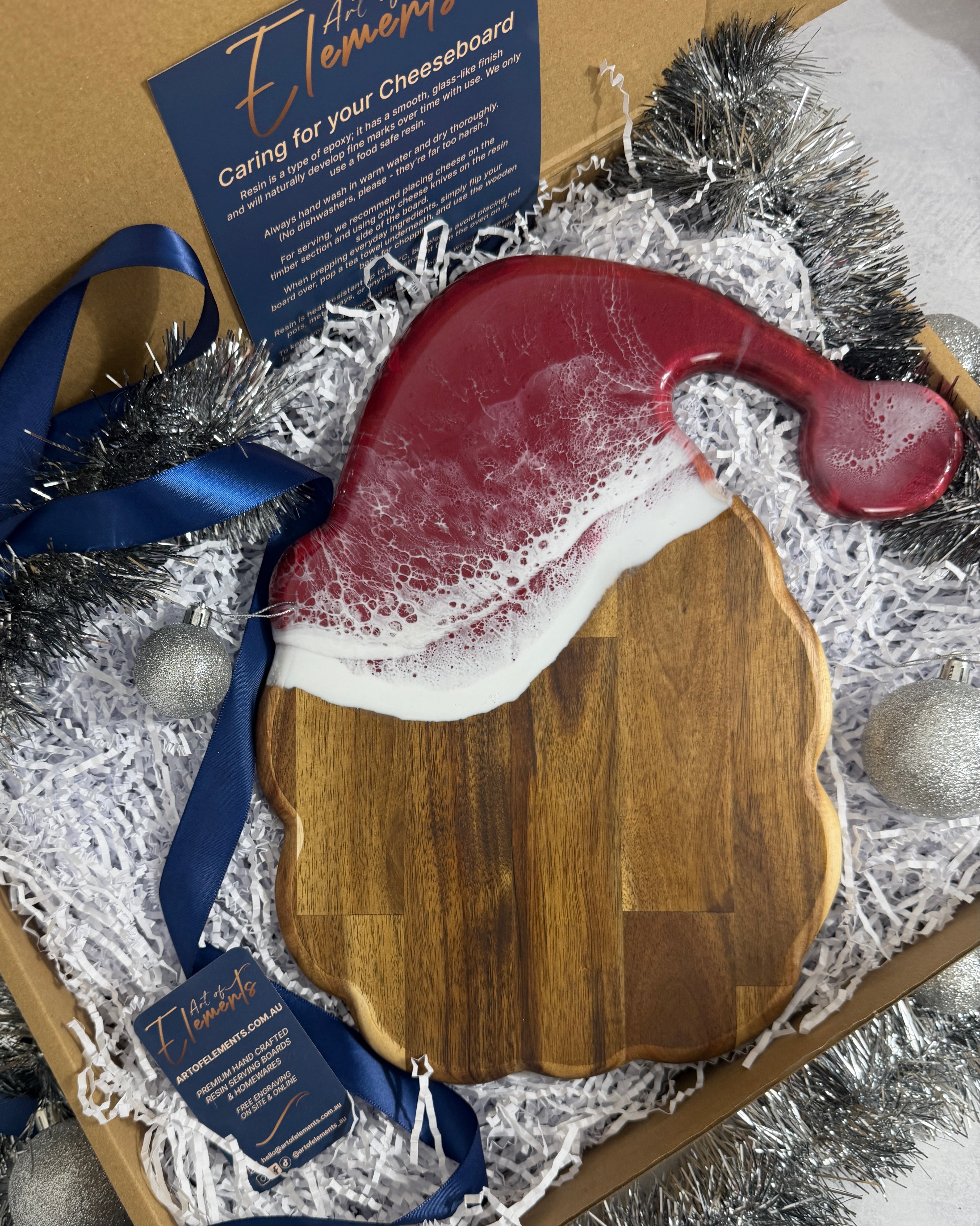A wooden cheeseboard shaped like a Santa hat, with red resin waves, presented in a gift box with silver tinsel, blue ribbons, and care instructions.