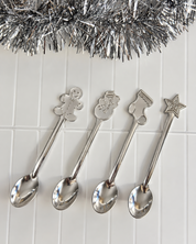 Four stainless steel spoons with holiday-themed handles, including a gingerbread man, snowman, stocking, and star, arranged on a white tiled surface with silver tinsel at the top.