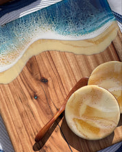 A styled flat-lay of a resin chopping board featuring an ocean wave design with blue and white foam over a beige sandy base, placed on a wooden board with two round coasters and a wooden knive.