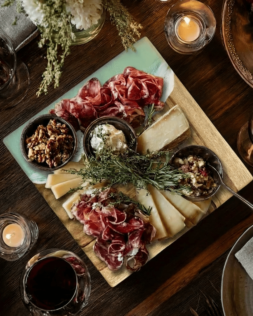 A mint green resin charcuterie board styled on a dark wooden table, featuring sliced cured meats, cheese wedges, crackers, nuts, and dips, surrounded by candles, a glass of red wine, and floral accents.
