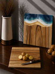 Camphor laurel timber square cutting board with potatoes and a knife on a wooden surface, with the same board with the resin front leaning against the kitchen wall