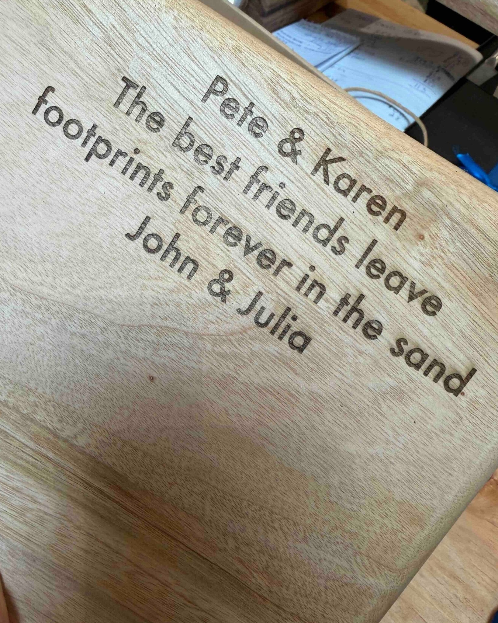 A close-up of a personalised wooden plaque engraved with the text 'Pete & Karen The best friends leave footprints forever in the sand John & Julia'.
