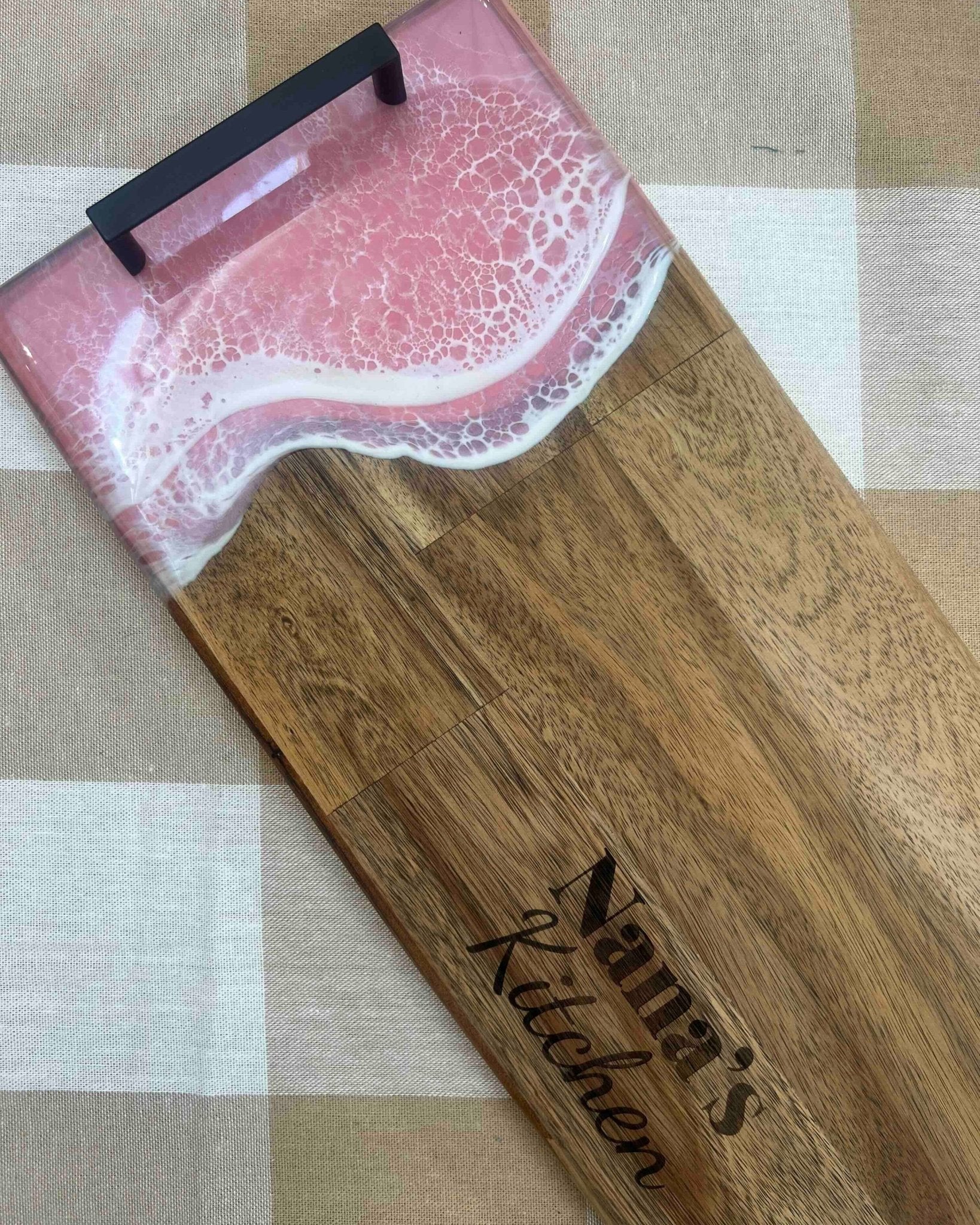 A wooden serving board or tray with a pink and white resin wave design at the top and the words 'Nana's Kitchen' engraved on the wood, placed on a beige and white checkered fabric surface.
