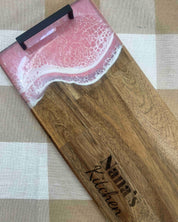 A wooden serving board or tray with a pink and white resin wave design at the top and the words 'Nana's Kitchen' engraved on the wood, placed on a beige and white checkered fabric surface.