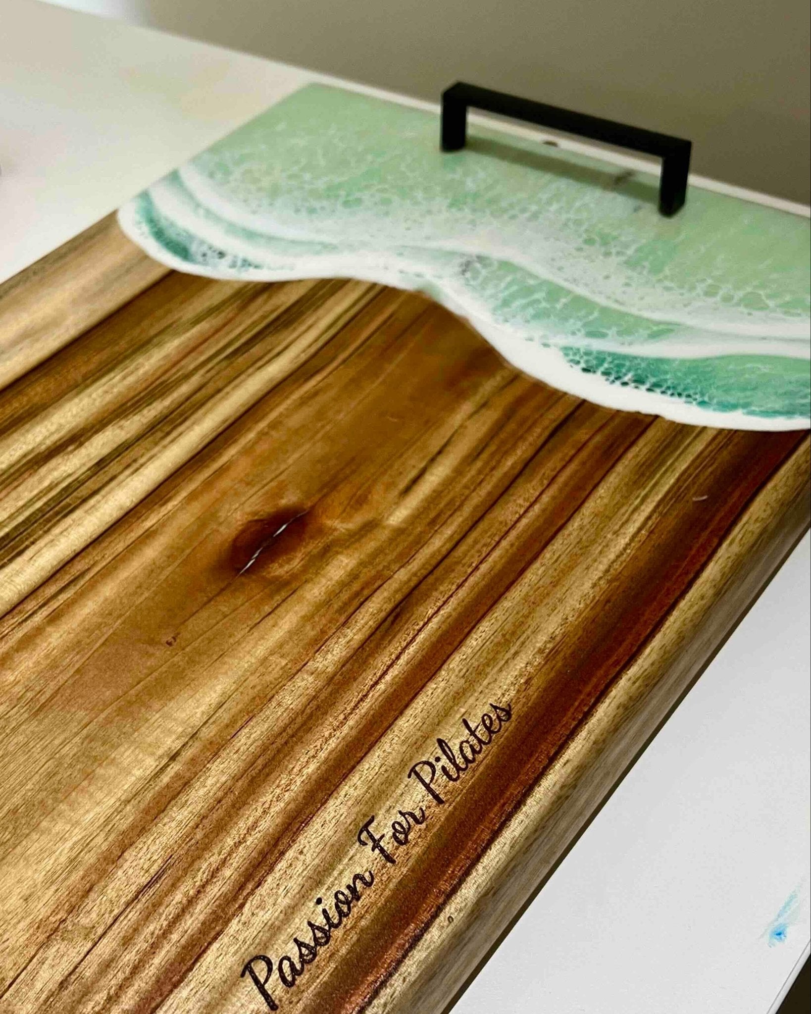 A wooden serving board with a wave-themed lid featuring green ocean waves and a black handle, resting on a white surface. The board is engraved with the text 'Passion For Plates'.