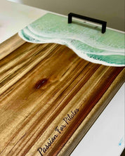 A wooden serving board with a wave-themed lid featuring green ocean waves and a black handle, resting on a white surface. The board is engraved with the text 'Passion For Plates'.