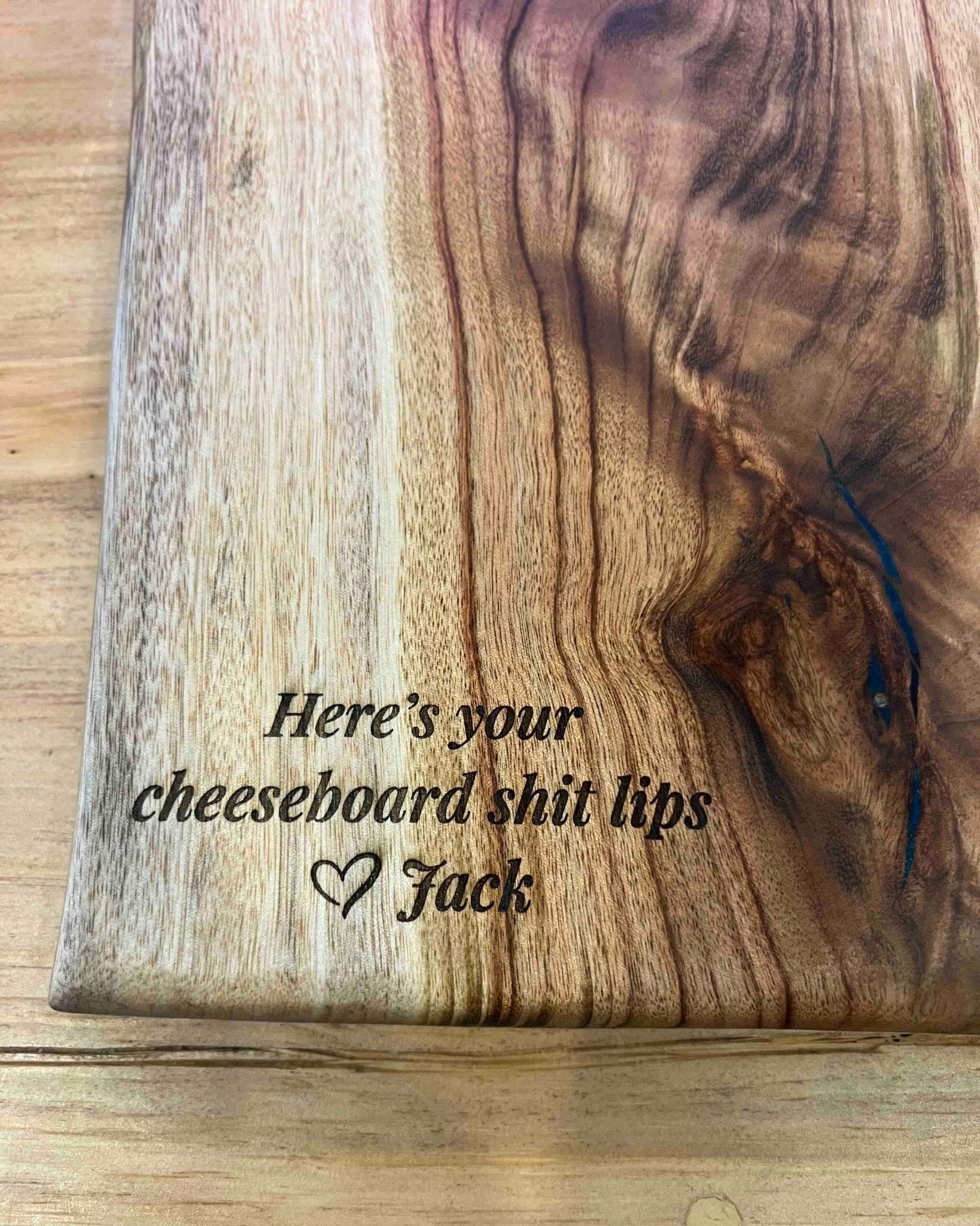 A close-up of a wooden cheeseboard with a natural grain and engraved text reading 'Here's your cheeseboard shit lips' and 'Jack' with a heart.
