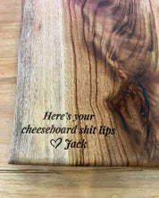 A close-up of a wooden cheeseboard with a natural grain and engraved text reading 'Here's your cheeseboard shit lips' and 'Jack' with a heart.