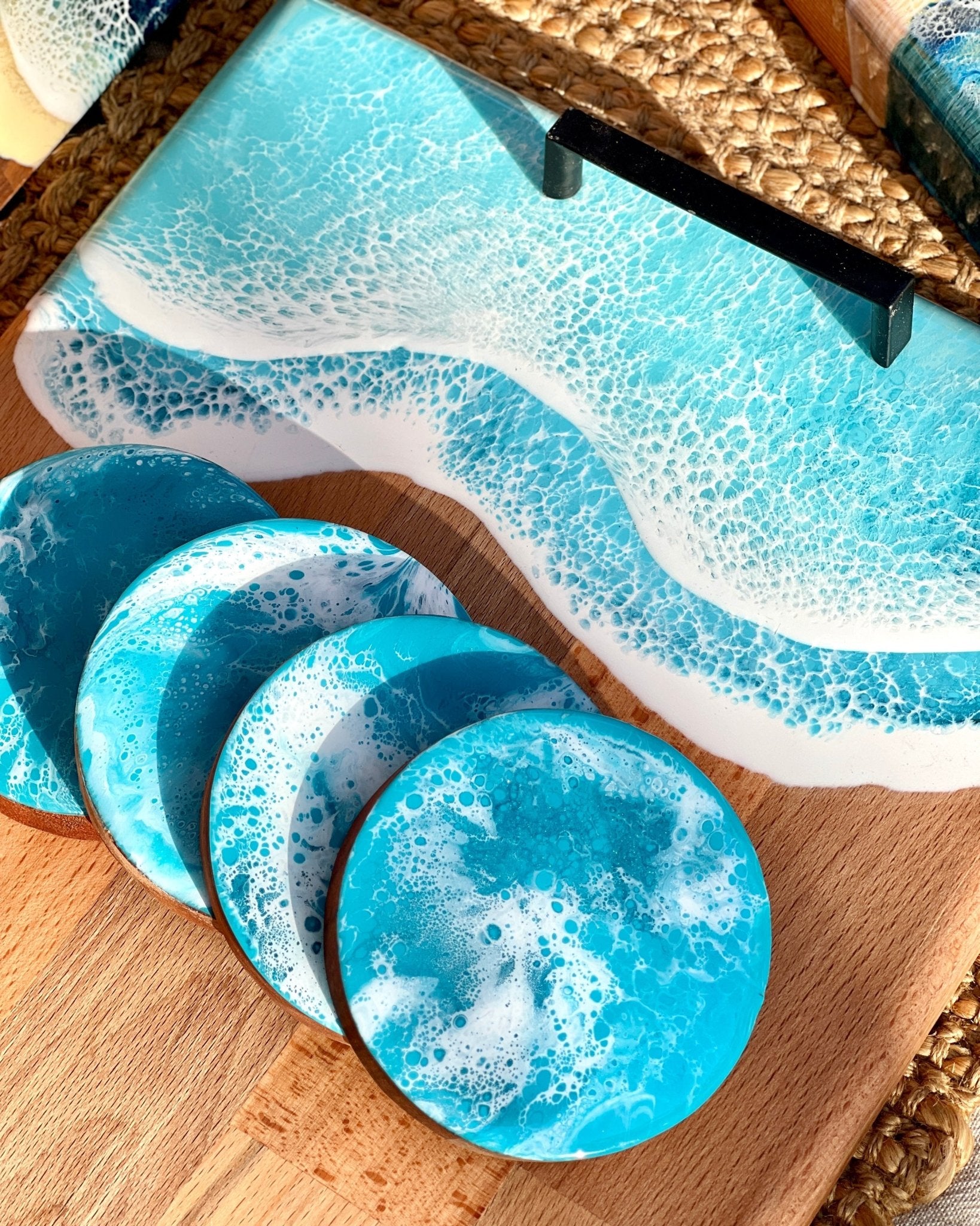 A set of round coasters with a pale blue and white ocean resin wave pattern, shown alongside a matching rectangular tray on a wooden board.