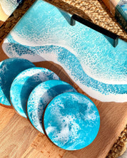 A set of round coasters with a pale blue and white ocean resin wave pattern, shown alongside a matching rectangular tray on a wooden board.
