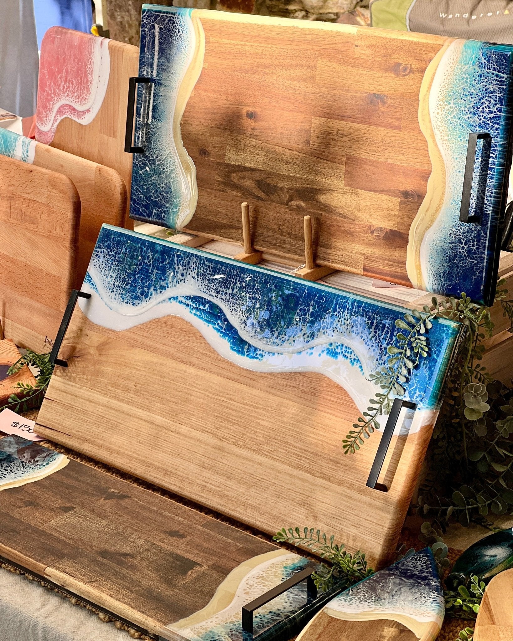 A set of wooden serving boards with hand-poured resin ocean wave designs in blue, white, with matte black metal handles