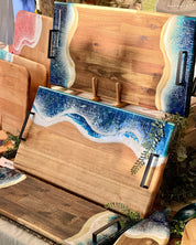 A set of wooden serving boards with hand-poured resin ocean wave designs in blue, white, with matte black metal handles