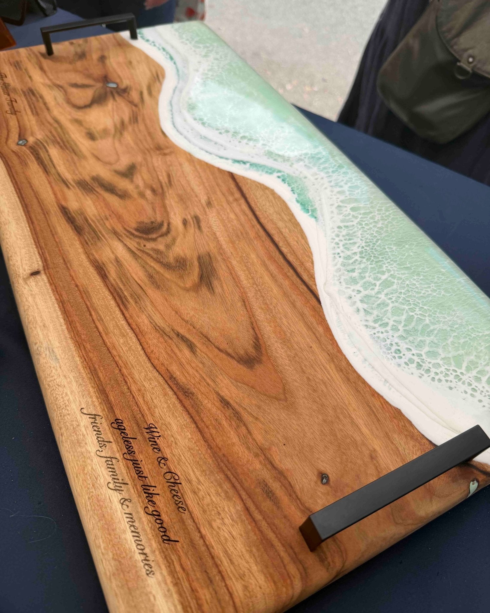 A rectangular wooden serving board with a wave design made from green and white resin, featuring a black metal handle and engraved text on a dark table.