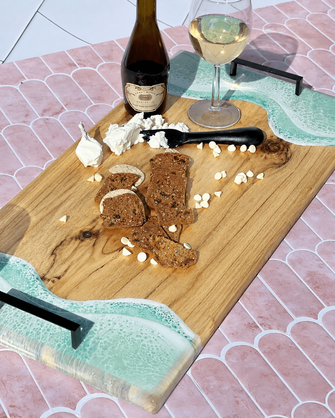 Wooden serving platter with metal handles and mint green resin waves with bread, cheese, and a bottle of wine on a tiled surface.