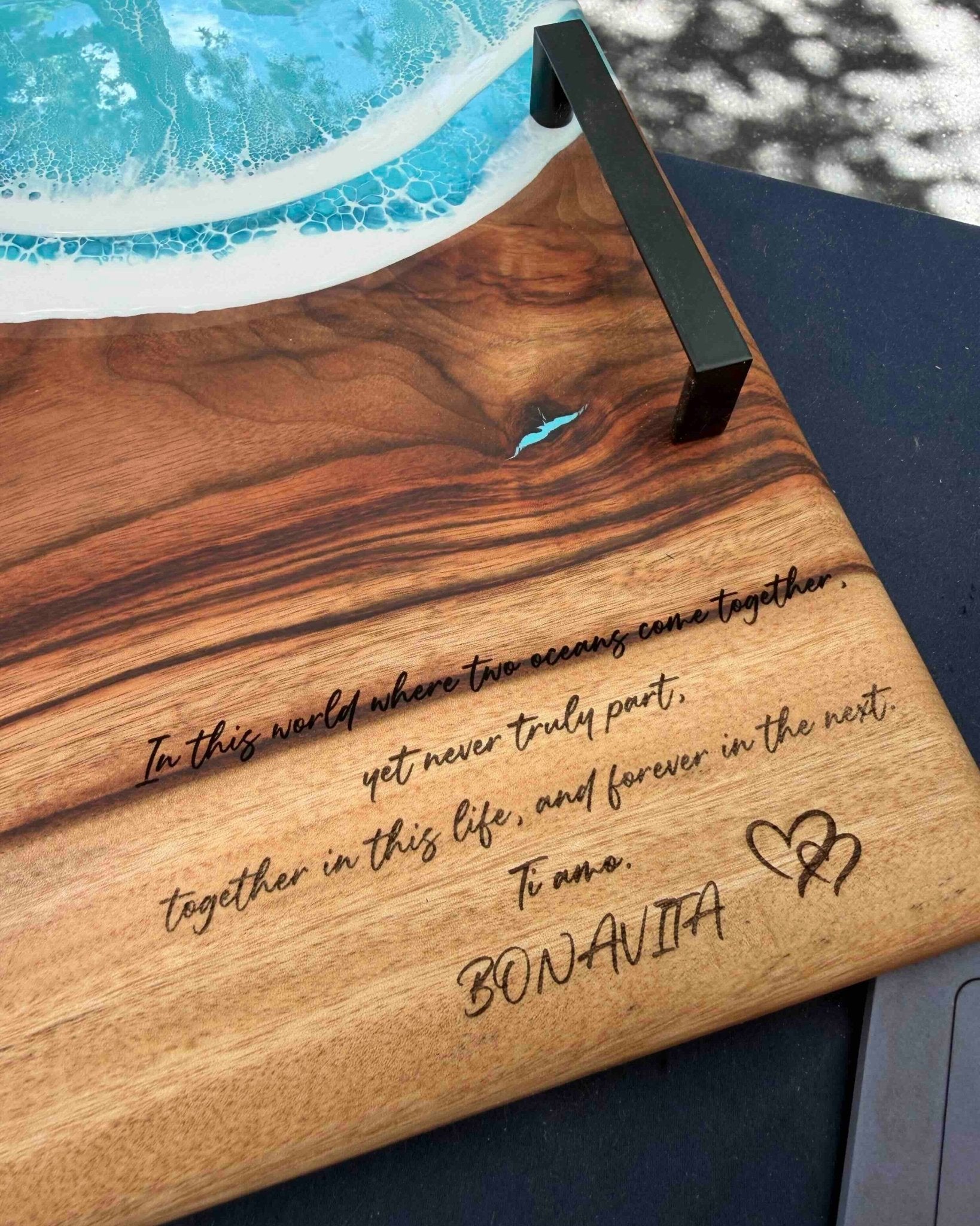 A close-up of a wooden serving board featuring a black metal handles and engraved text that reads, 'In this world where two means can co-exist, yet never truly part, together in this life, and forever in the next. Ti Amo"