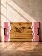Wooden cutting board with pink resin edges, black metal handles on a wooden surface