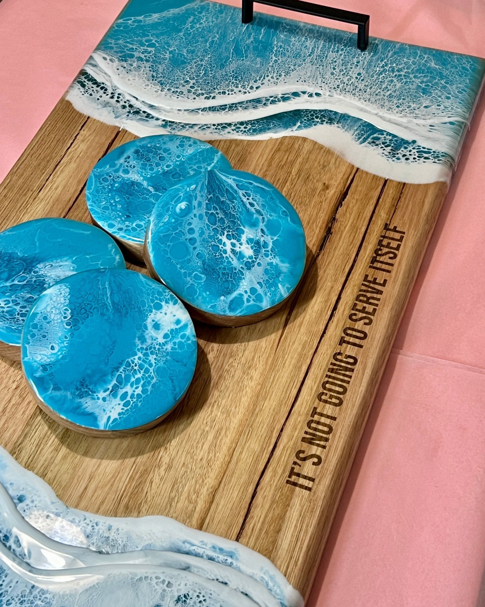 Wooden cutting board with turquoise  resin coasters and a pink background, with the engraved" It's not going to serve itself" 