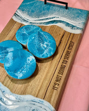 Wooden cutting board with turquoise  resin coasters and a pink background, with the engraved" It's not going to serve itself" 