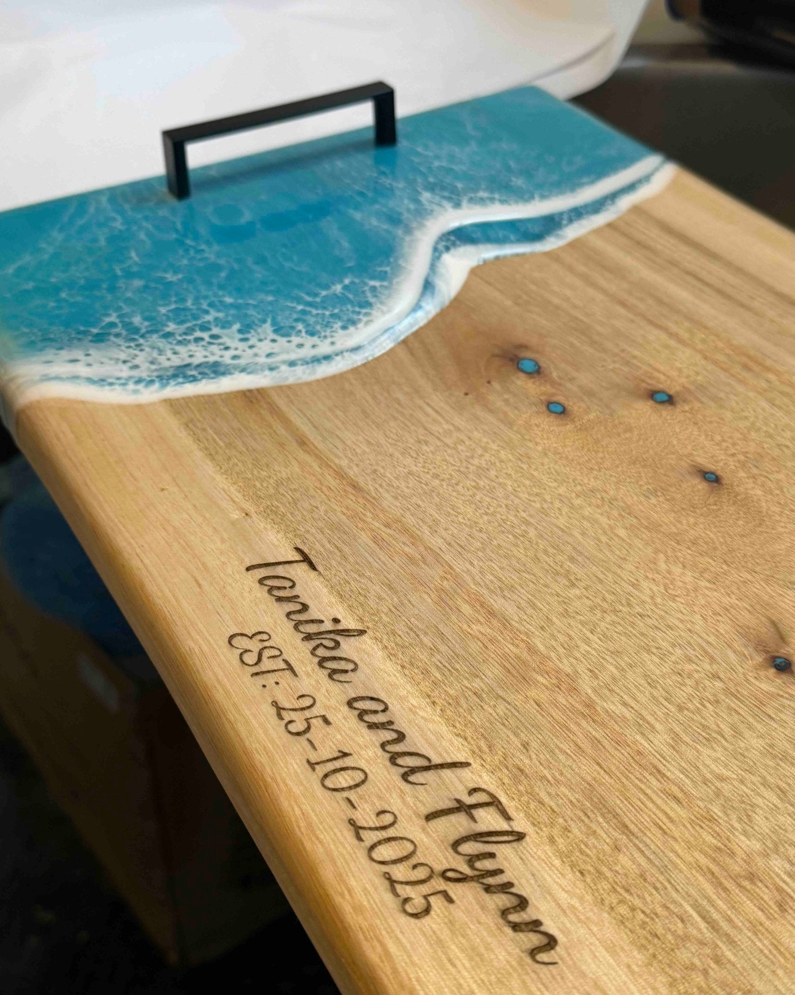 A personalised wooden serving board with a turquoise resin ocean wave design and a black metal handles. The board is engraved with the names 'Tanisha and Flynn' and the date 'EST 25-10-2025"