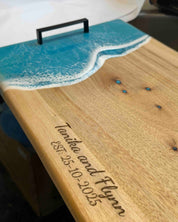 A personalised wooden serving board with a turquoise resin ocean wave design and a black metal handles. The board is engraved with the names 'Tanisha and Flynn' and the date 'EST 25-10-2025"
